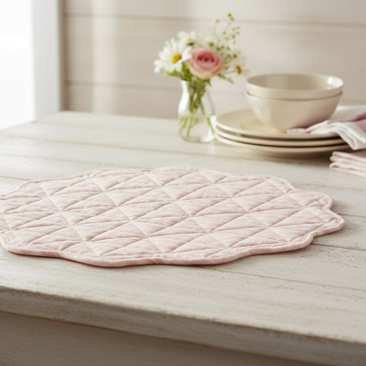 Pink quilted placemat on a wooden surface with a bowl and flowers in the background.