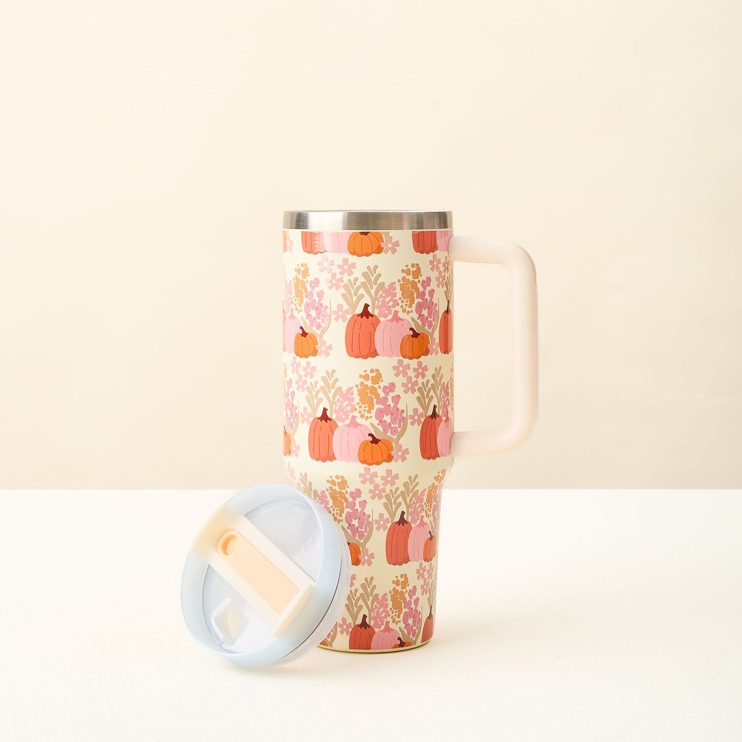 Cream colored tumbler with handle and pink and orange foliage and pumpkins with a lid sitting to the side.