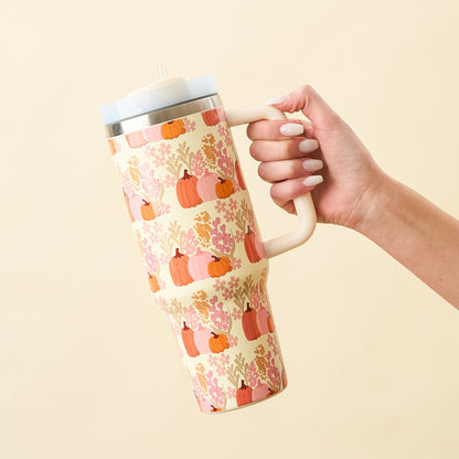 A hand holds a cream colored tumbler with pink and green foliage and pumpkins.