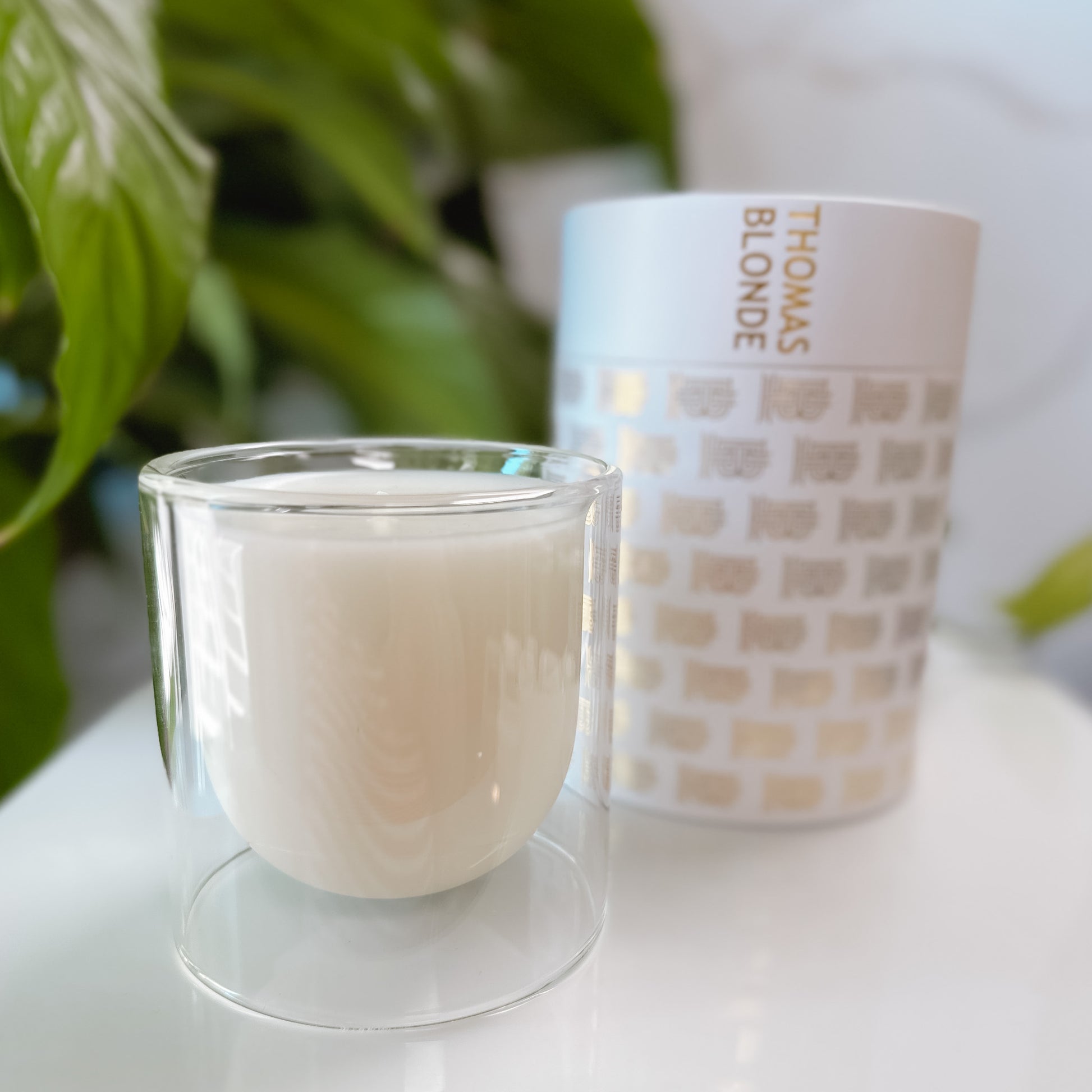 Thomas Blonde candle in a clear glass jar with it's packaging on a white tray with foliage in the background.