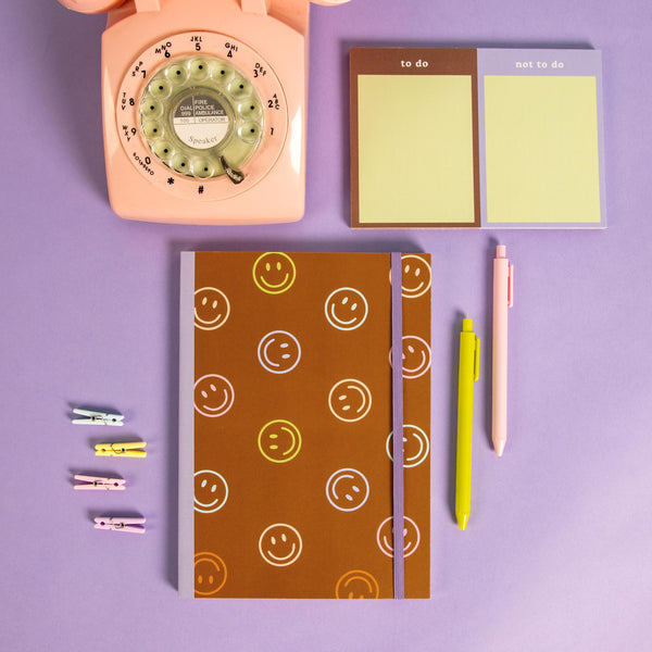 smiley notebook on a desk with a phone, clips, and pens.