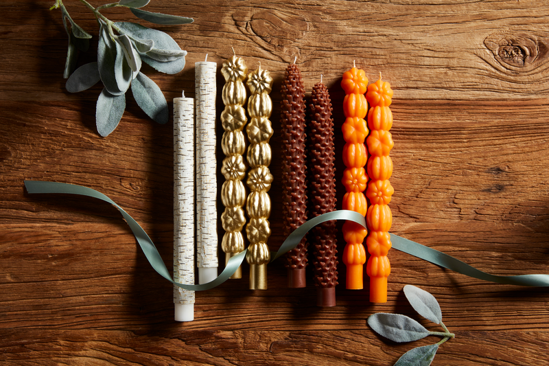 4 styles of taper candles laying on a wooden table with a piece of ribbon and sprigs of greenery.
