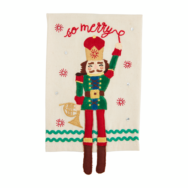 off-white towel with nutcracker holding a horn appliqued on it and so merry embroidered across the top.