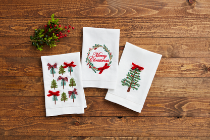 3 white towels with christmas embroidery on them arranged on a wooden background with a sprig of greenery.