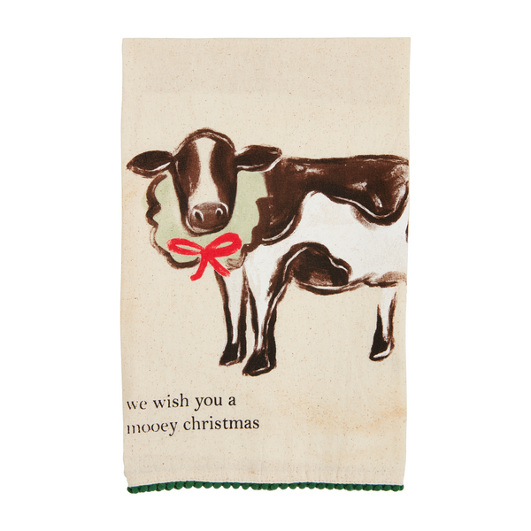 off-white towel with green pom trim with a cow wearing a wreath around its neck and "we wish you a mooey christmas" printed on it.