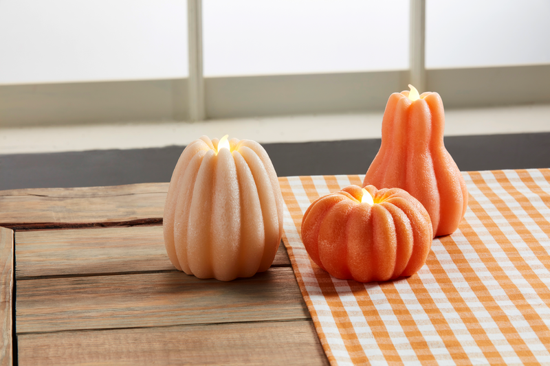 3 styles of lit pumpkin candles arranged on a wooden table with an orange checkered runner.