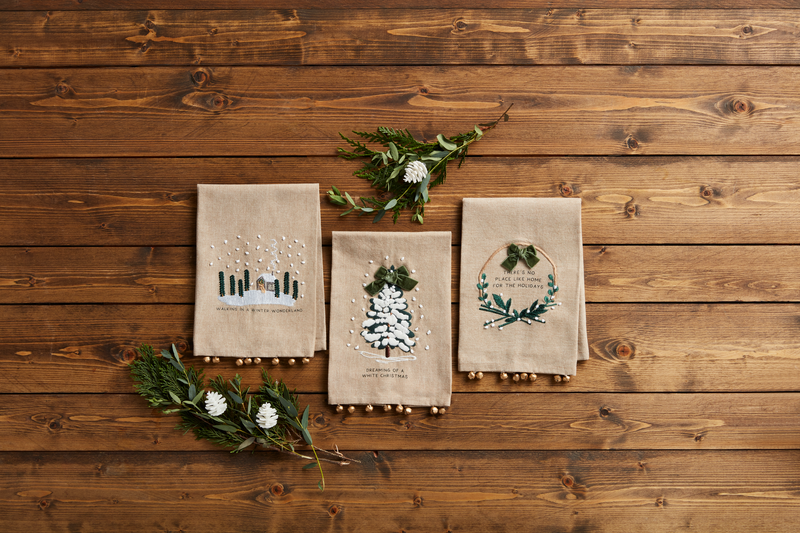 3 styles of embroidered christmas towels arranged on a wood slap background with sprigs of greenery.