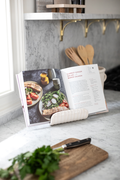 ceramic cookbook holder on a kitchen countertop with an open cookbook in it and a cutting board with greens and a kinife on it.