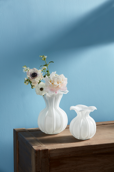 2 sizes of white glass ruffle vases, large one is filled with flowers, set on a wooden table in front of a blue wall.
