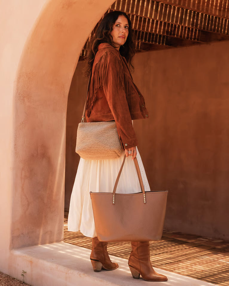 Woman in a brown suede jacket and white skirt holding a beige crossbody and tote against a warm-toned wall.