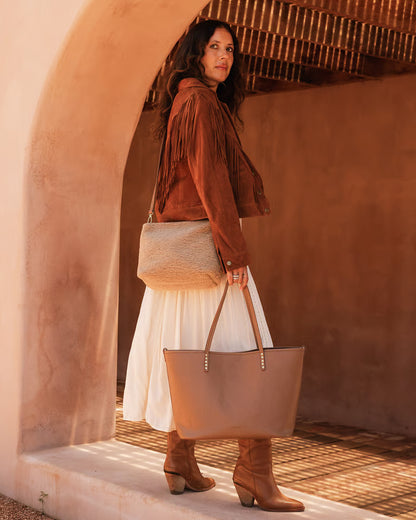 Woman in a brown suede jacket and white skirt holding a beige crossbody and tote against a warm-toned wall.