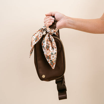 hand holding mocha brown all your need bag by the strap on a blush pink background.