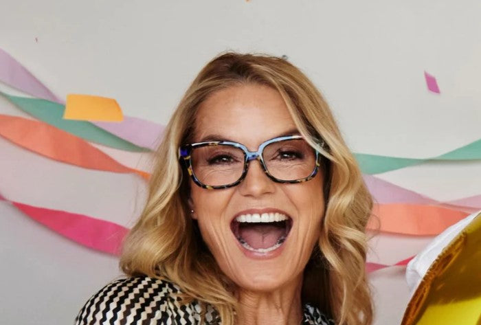 a woman modeling the Peepfetti Tortoise Impromptu Blue Light Reading Glasses while standing in front of a wall with streamers and falling confetti