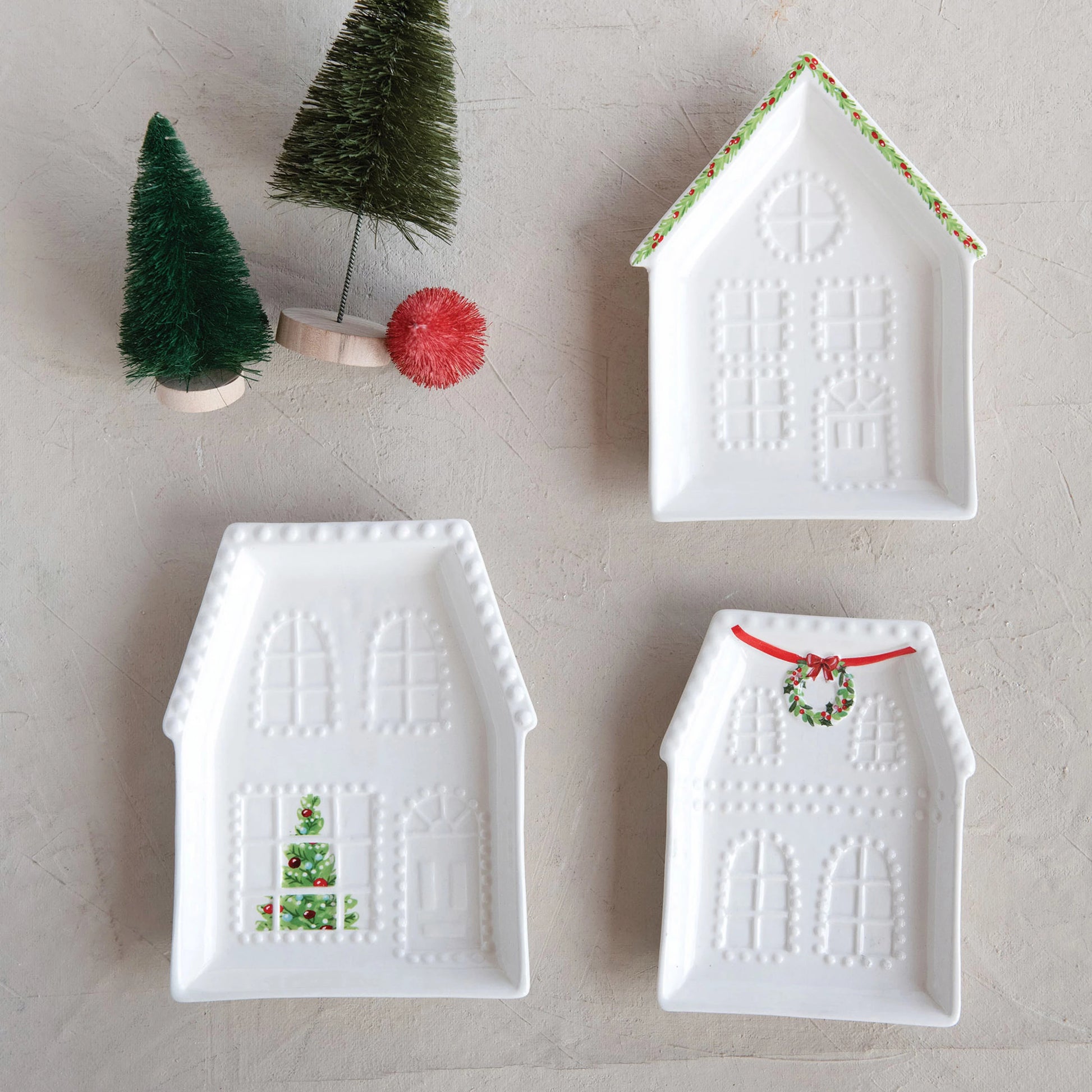 Three house shaped little plates arranged on a plaster surface with a few bottle brush trees.