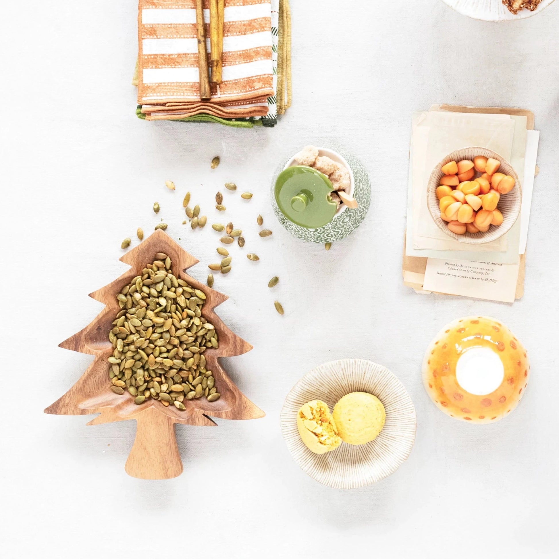top view of the acacia wood christmas tree shaped bowl filled with pine nuts and displayed next to striped towels, gold flatware, bowls of dip, mug, and plate of bread on a white surface