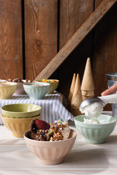 several assorted ice cream bowls arranged on a wooden table, some stacked, some filled with ice cream, others filled with toppings.