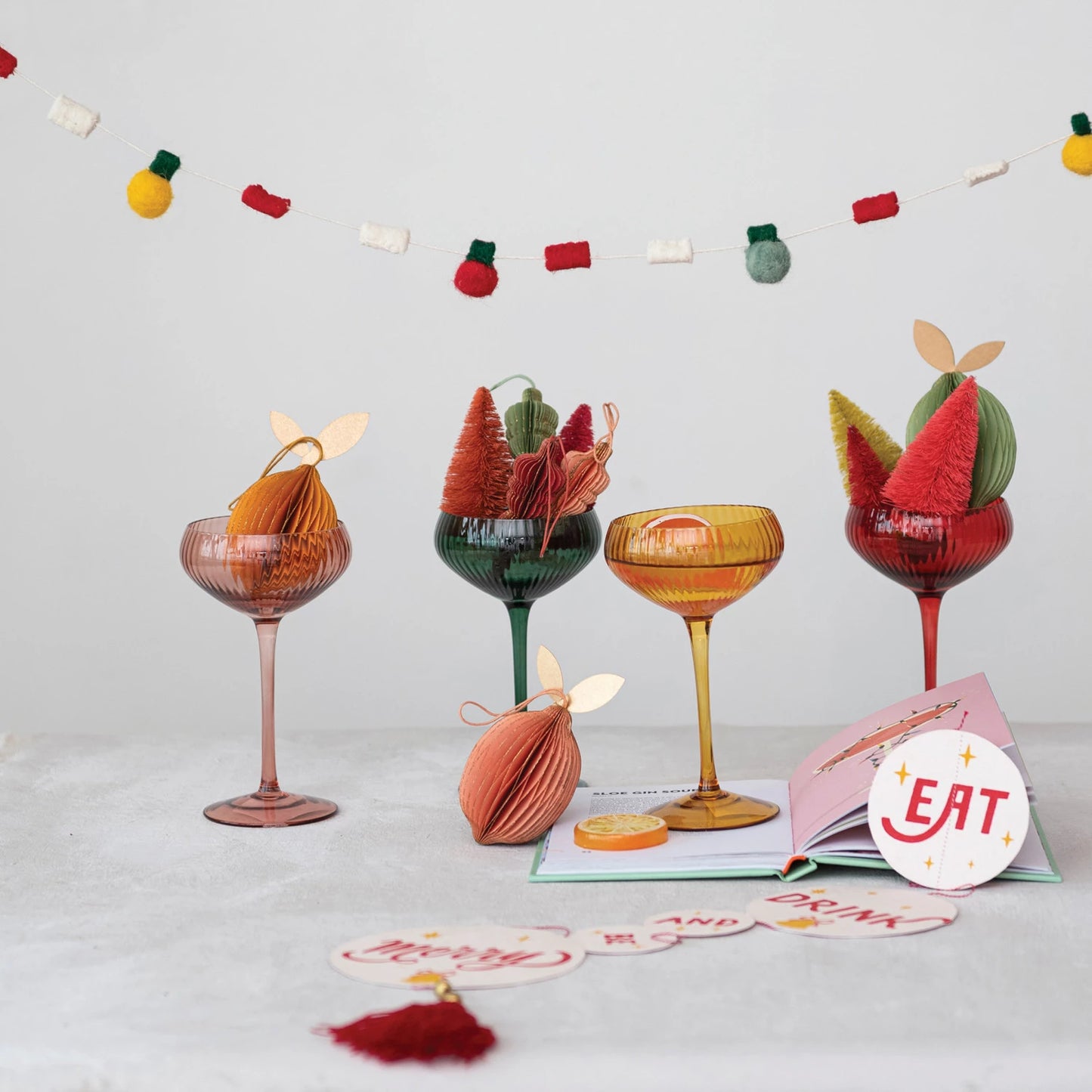 all four stemmed champagne coup glasses displayed with paper ornaments and holiday decorations and book with  holiday garland hanging in the background