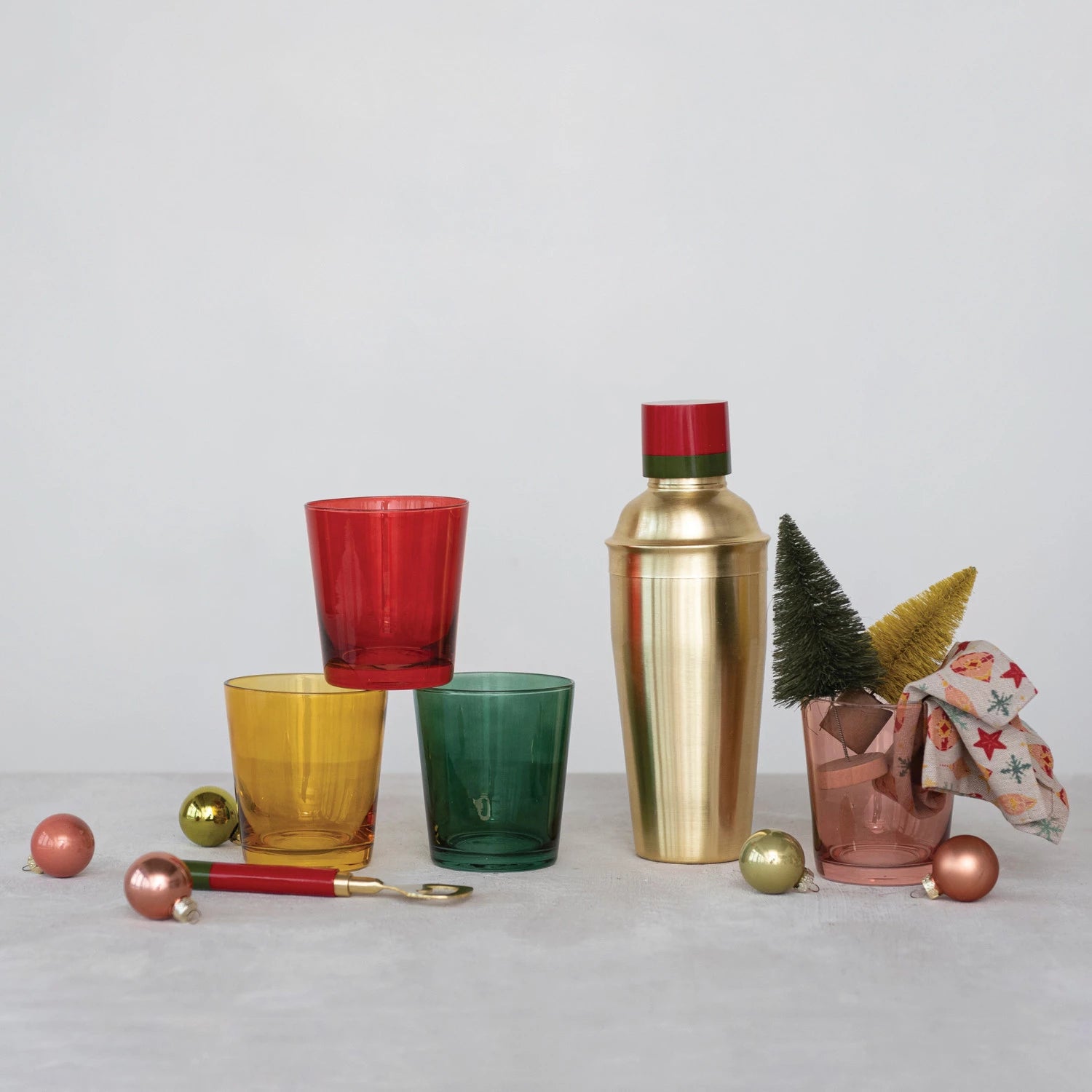 all four colors of low ball drinking glasses displayed next to a gold cocktail shaker, small ornaments, bottle opener, and small bottle brush trees against a light gray background