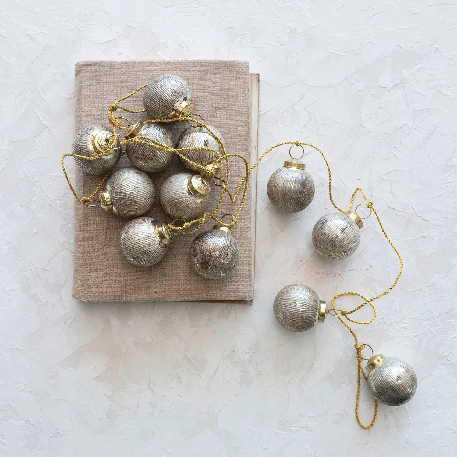 glass ball ornament garland with gold cord displayed on a linen covered book on a white plaster background