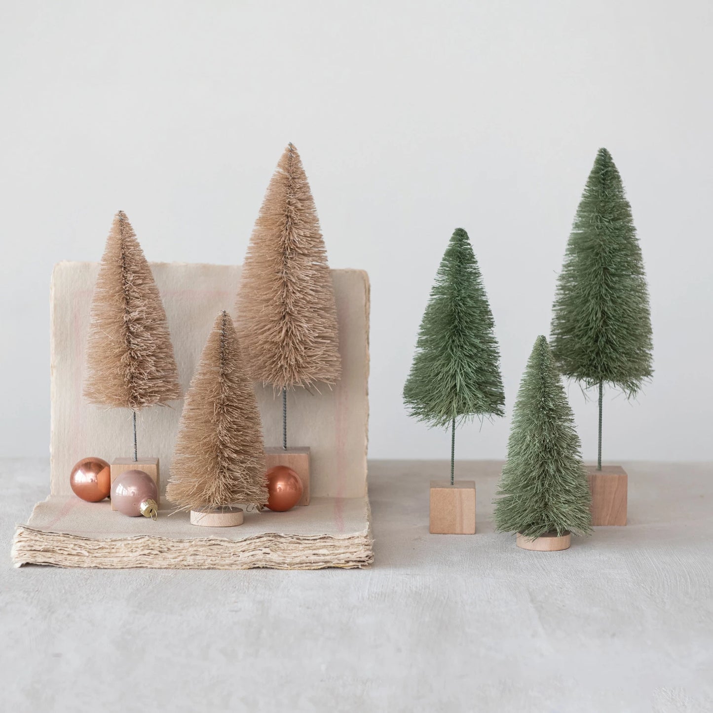 all three sizes of sage sisal bottle brush trees displayed next to cream ones with neutral small ornaments on a stack of unbleached paper