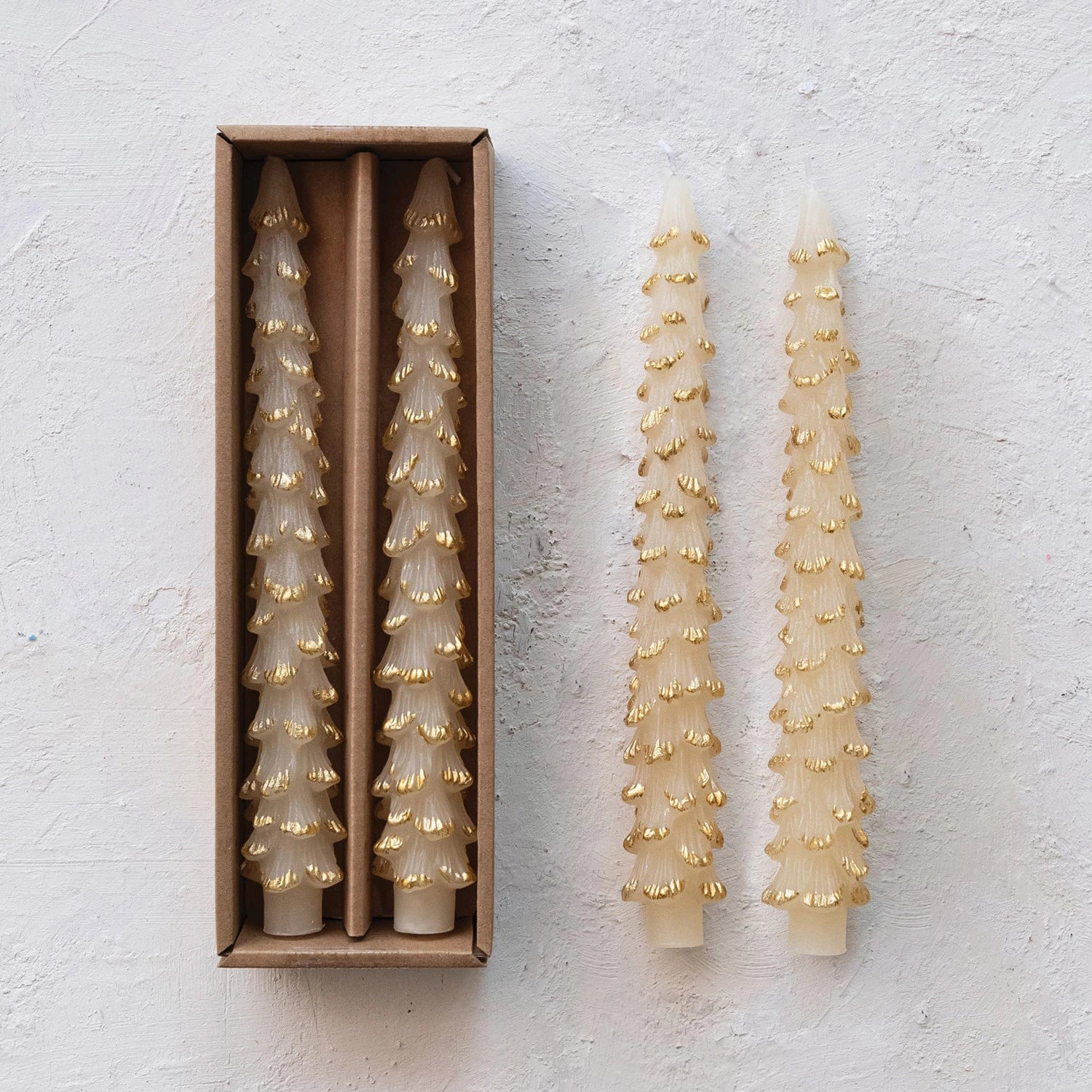 2 ivory tree shaped candles with gold accents laying on a plaster surface next to 2 other candles in their box packaging.