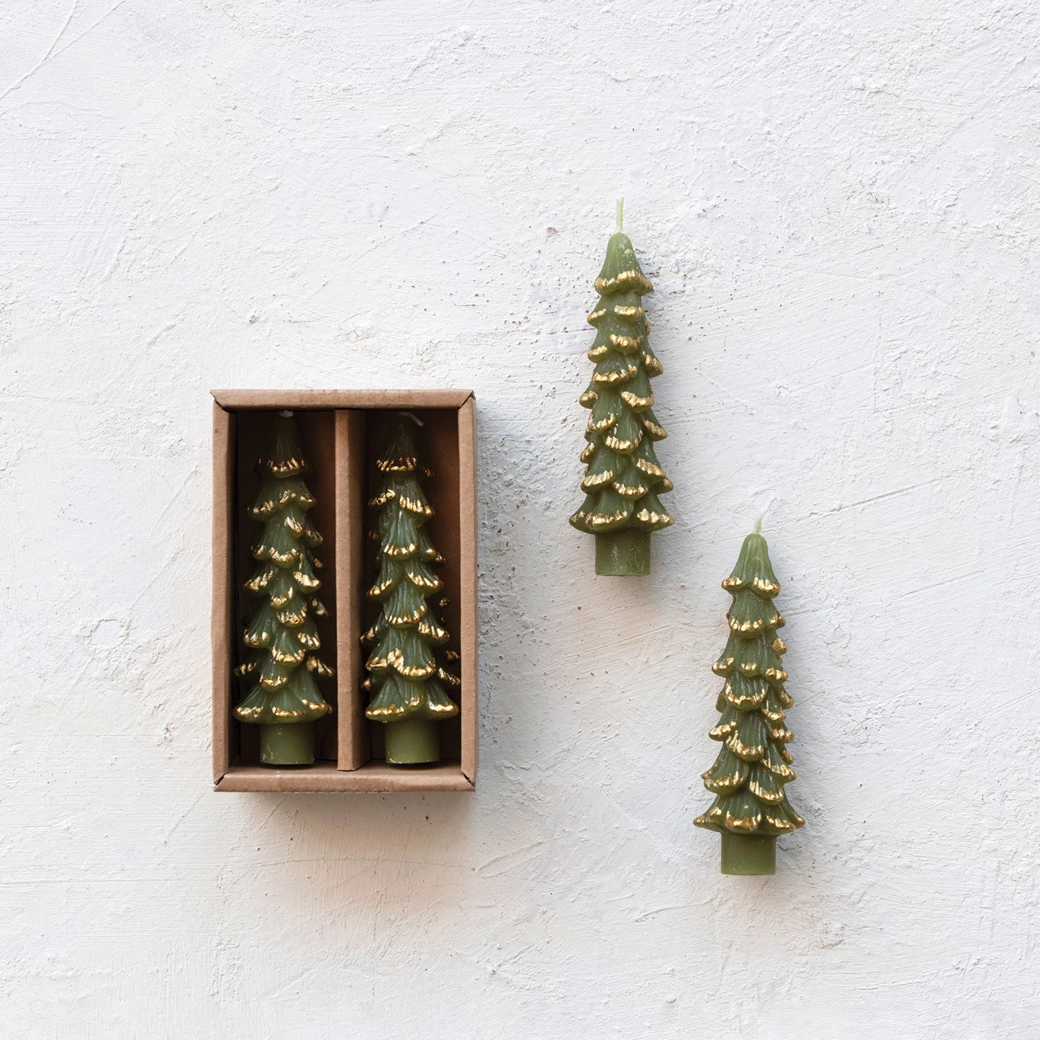 green tree shaped candles laying on a plaster surface with 2 other candles in their box packaging.