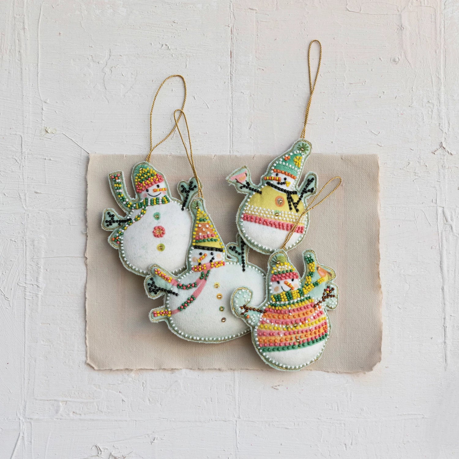 assorted velvet snowman ornaments embroidered with colorful glass beads arranged on a piece of paper set on a plaster surface.