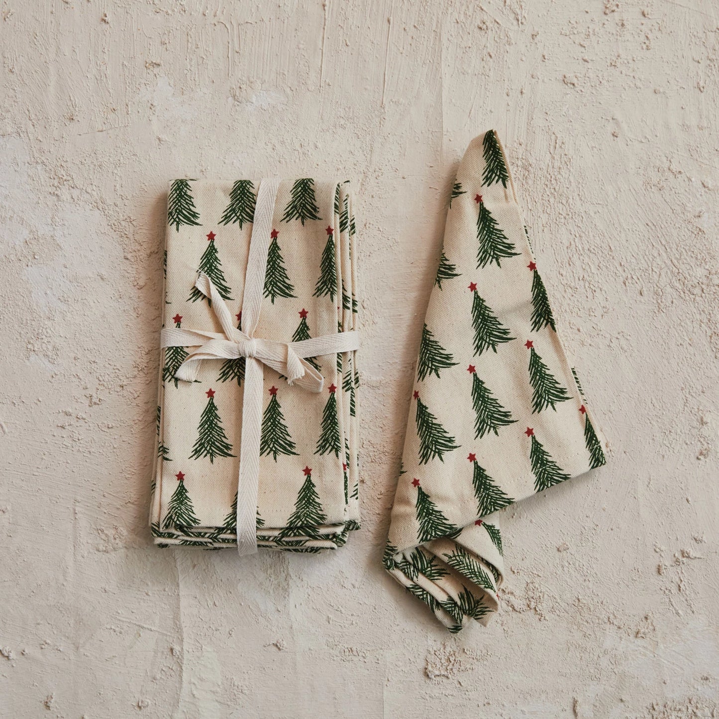 christmas tree napkin set tied together with a ribbon and a christmas tree napkins draped next to it on a plaster background.