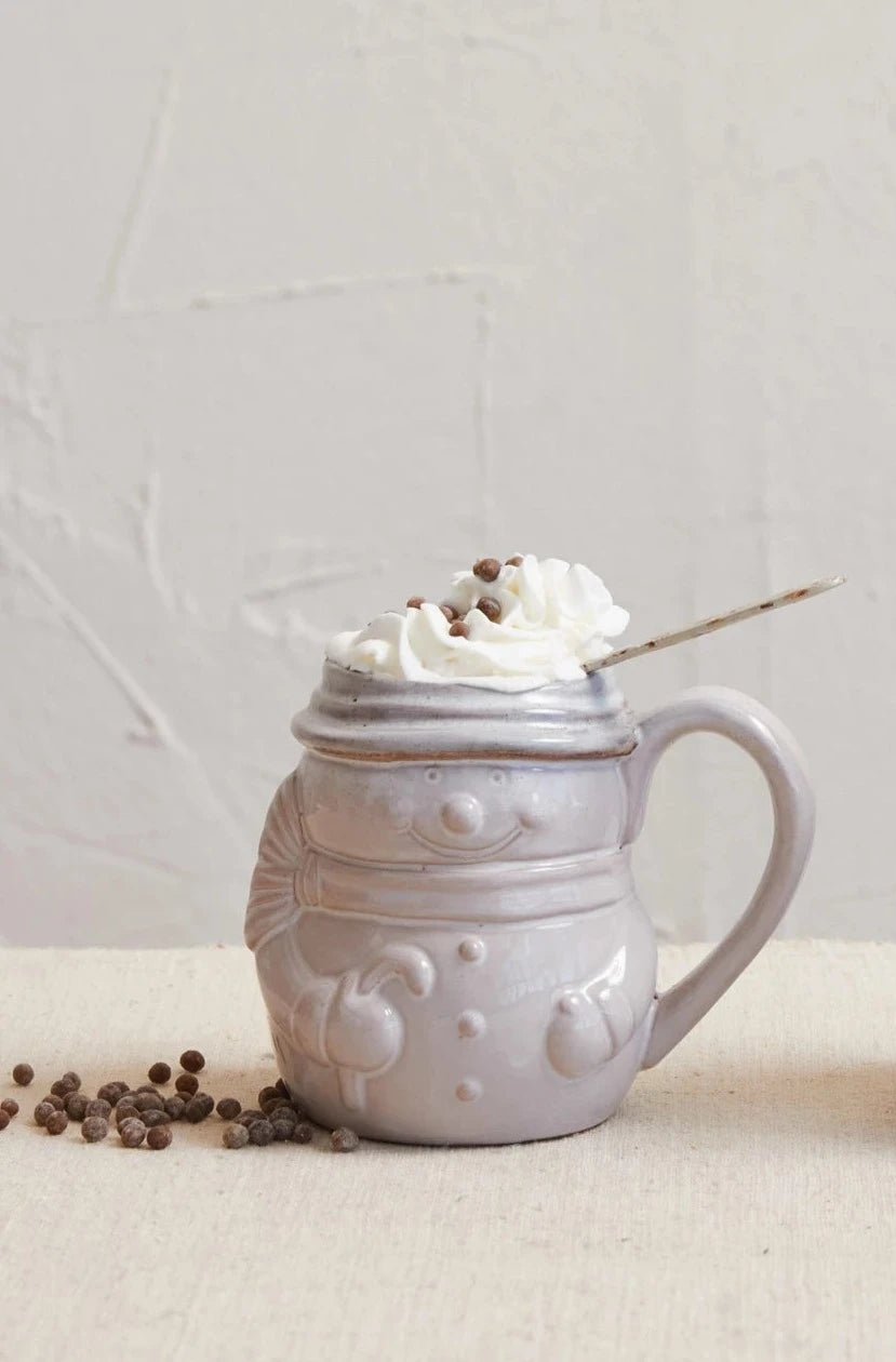 snowman mug filled with beverage and topped with whipping cream and spices arranged on a table with a plaster background.