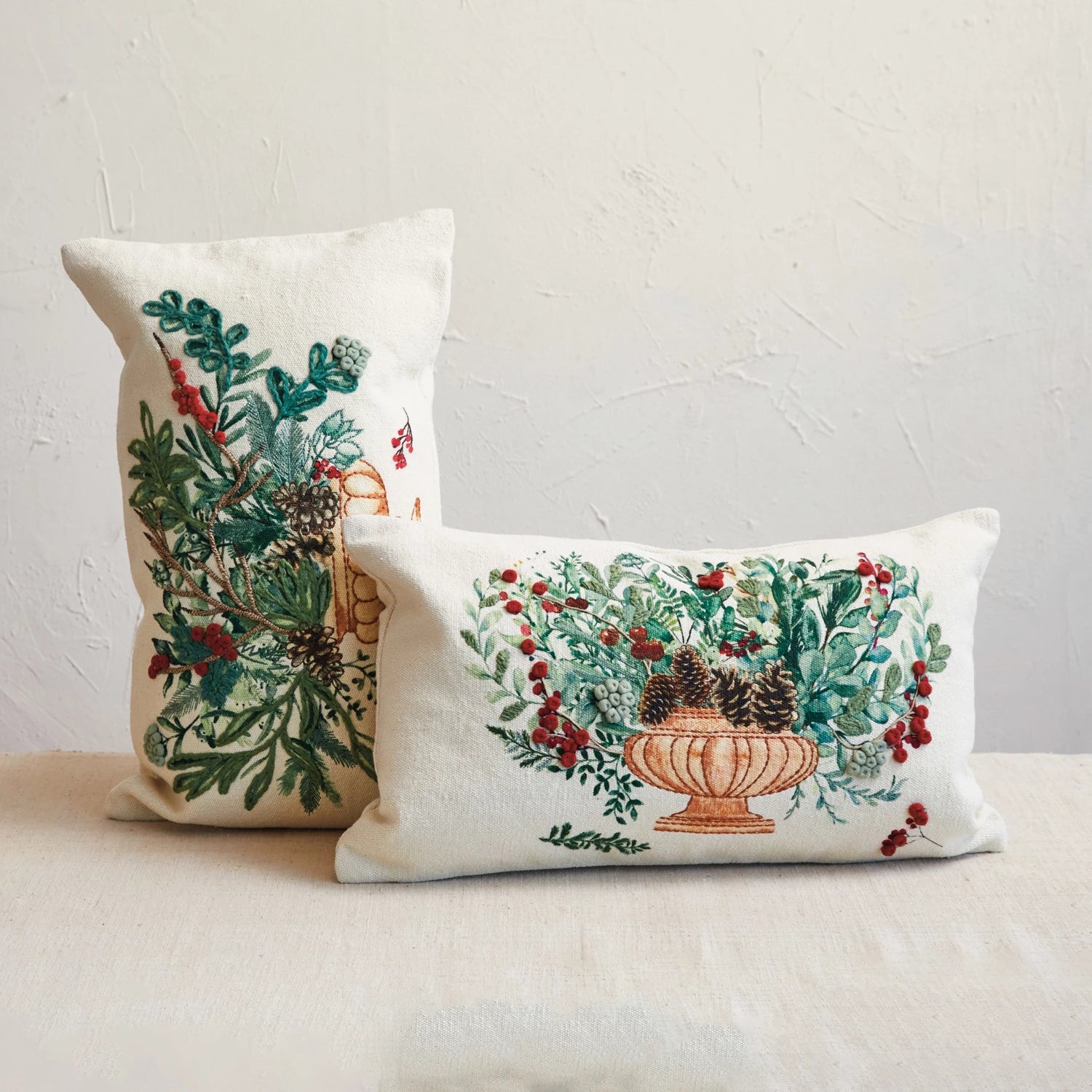 2 winter botanical pillows arranged on a table in front of a plaster background.