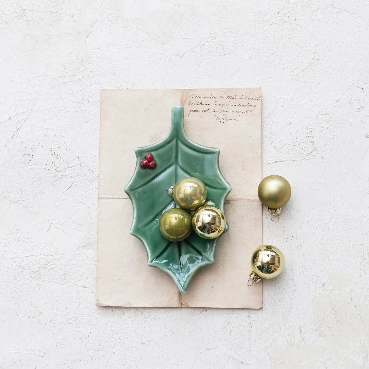 holly leaf plate set on a stack of off-white papers with gold ball ornaments arranged on it and next to it.