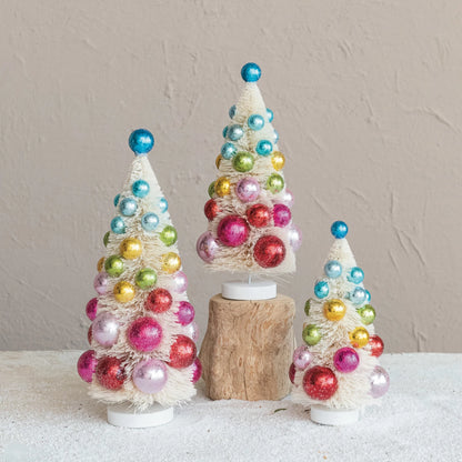 3 sizes of off-white sisal trees with colorful ornaments arranged on a table, the medium sized tree is set on a block of wood.