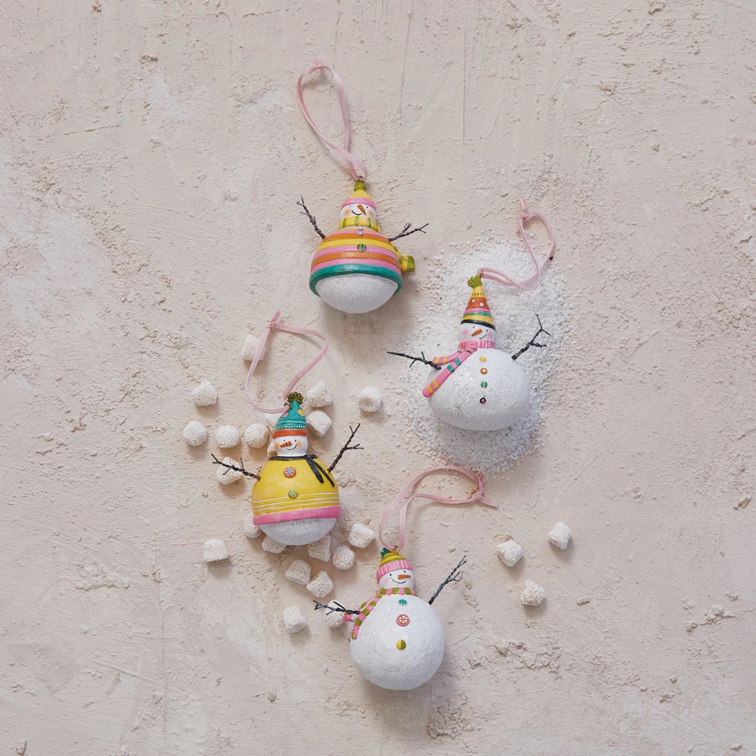 4 styles of snowman ornaments arranged on a plaster surface that is sprinkled with artificial snow and marshmallows.