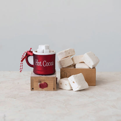 red glass mug ornament set on a block of wood with a stack of marshmallows next to it.