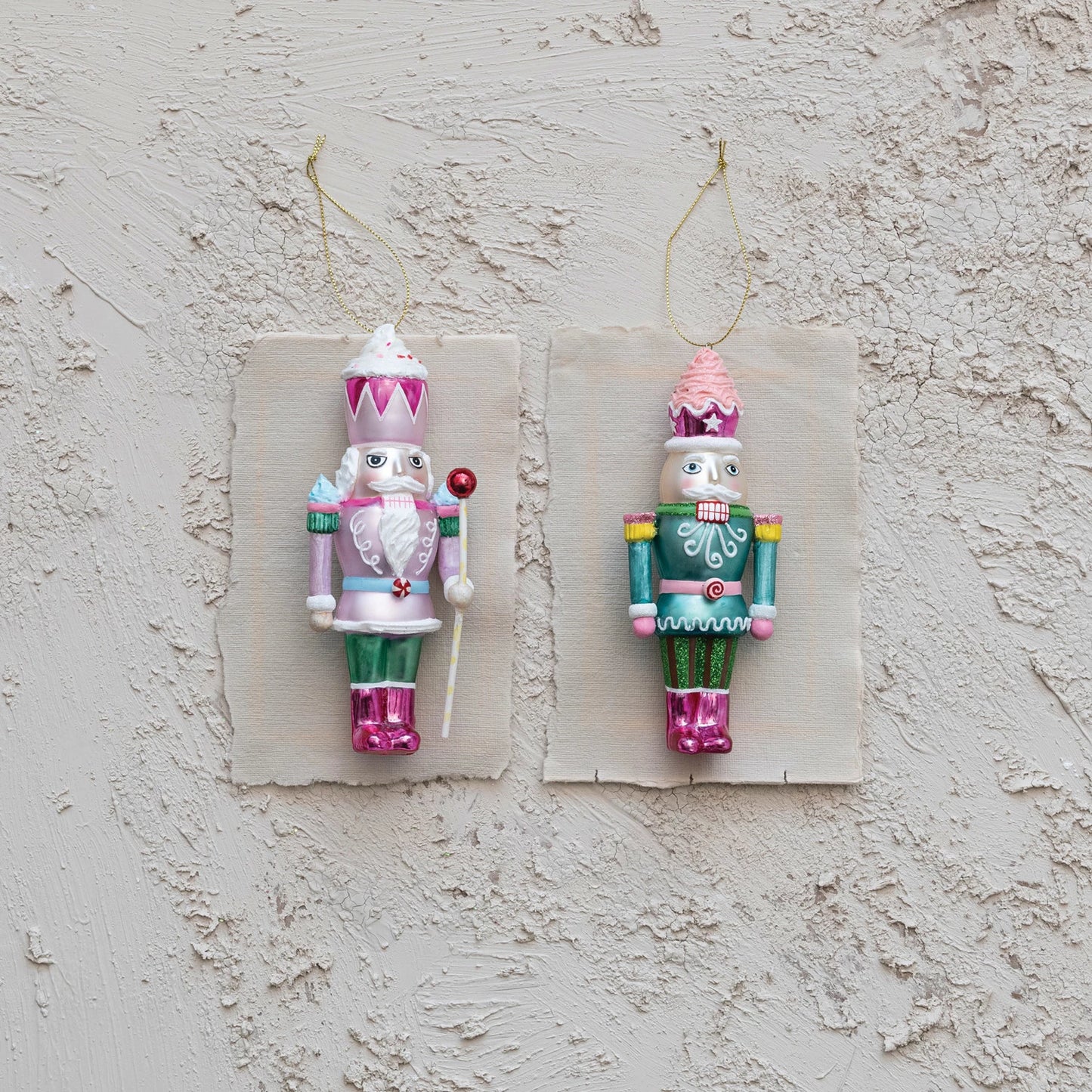 Two styles of pink and green glass nutcracker ornaments laying on bits of paper arranged on a plaster background.