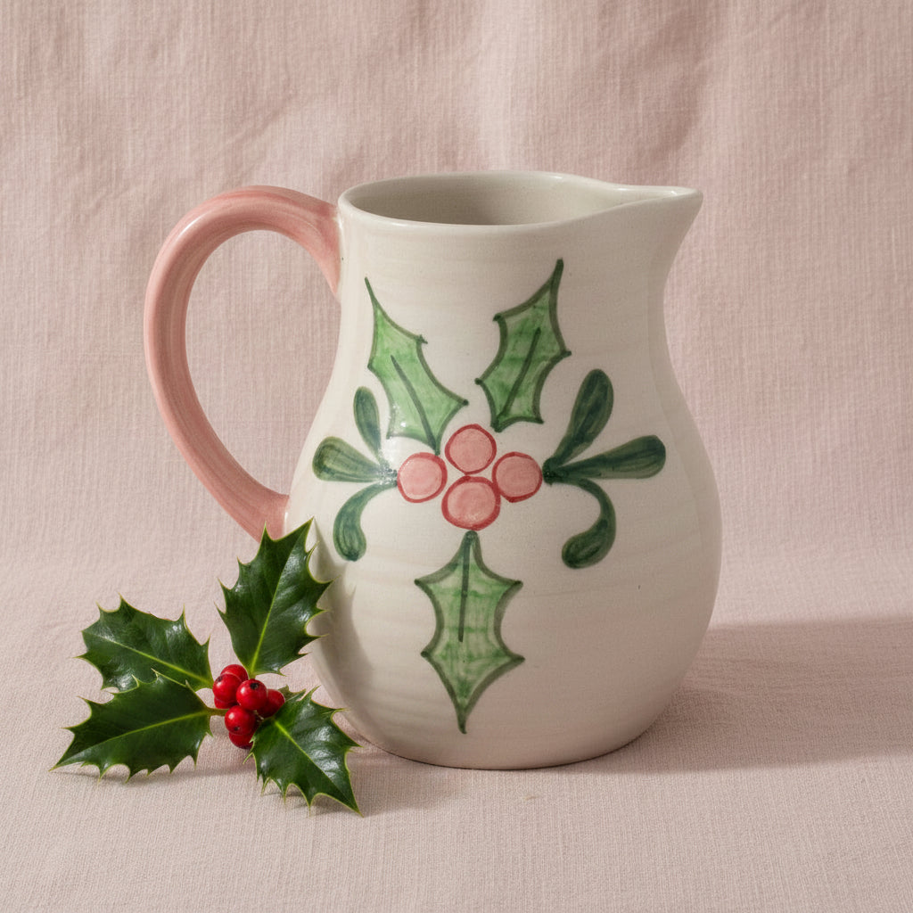 Ceramic pitcher with holly design shown on a pink background with a sprig of holly next to it