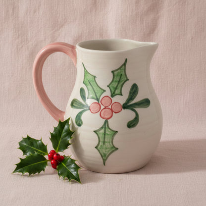 Ceramic pitcher with holly design shown on a pink background with a sprig of holly next to it
