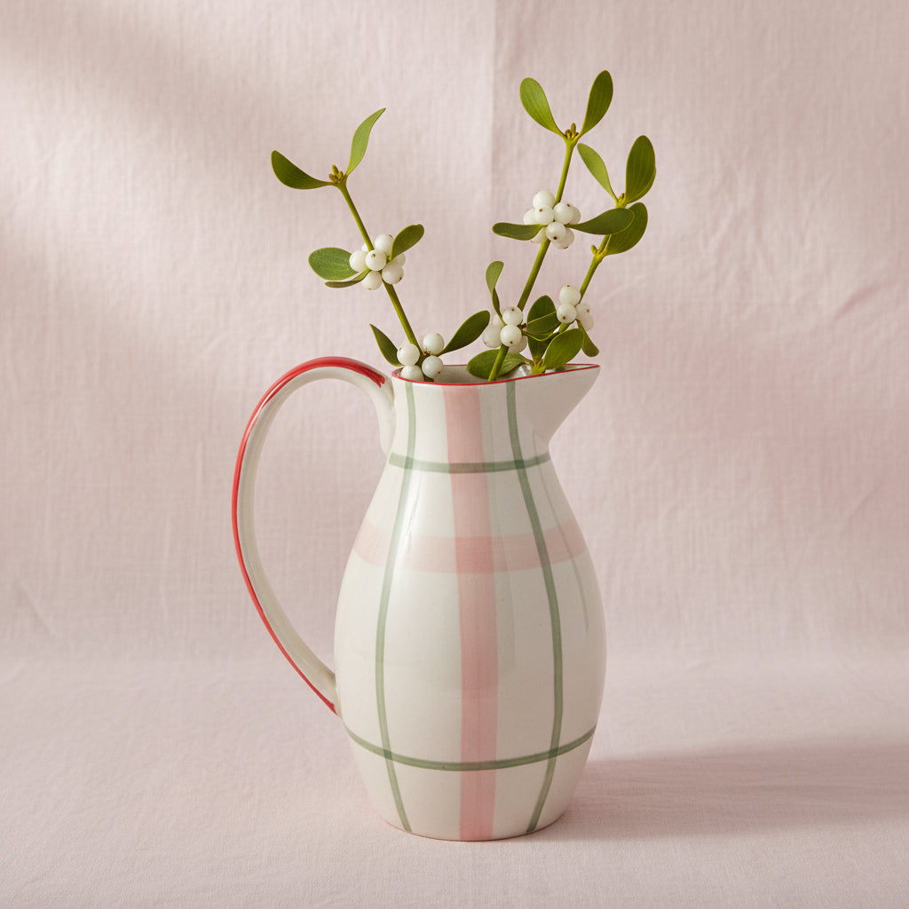 A ceramic pitcher with a rounded body, a curved handle, and spout, decorated with a plaid pattern in soft pink and green shown on a pale pink background with mistletoe stems in it