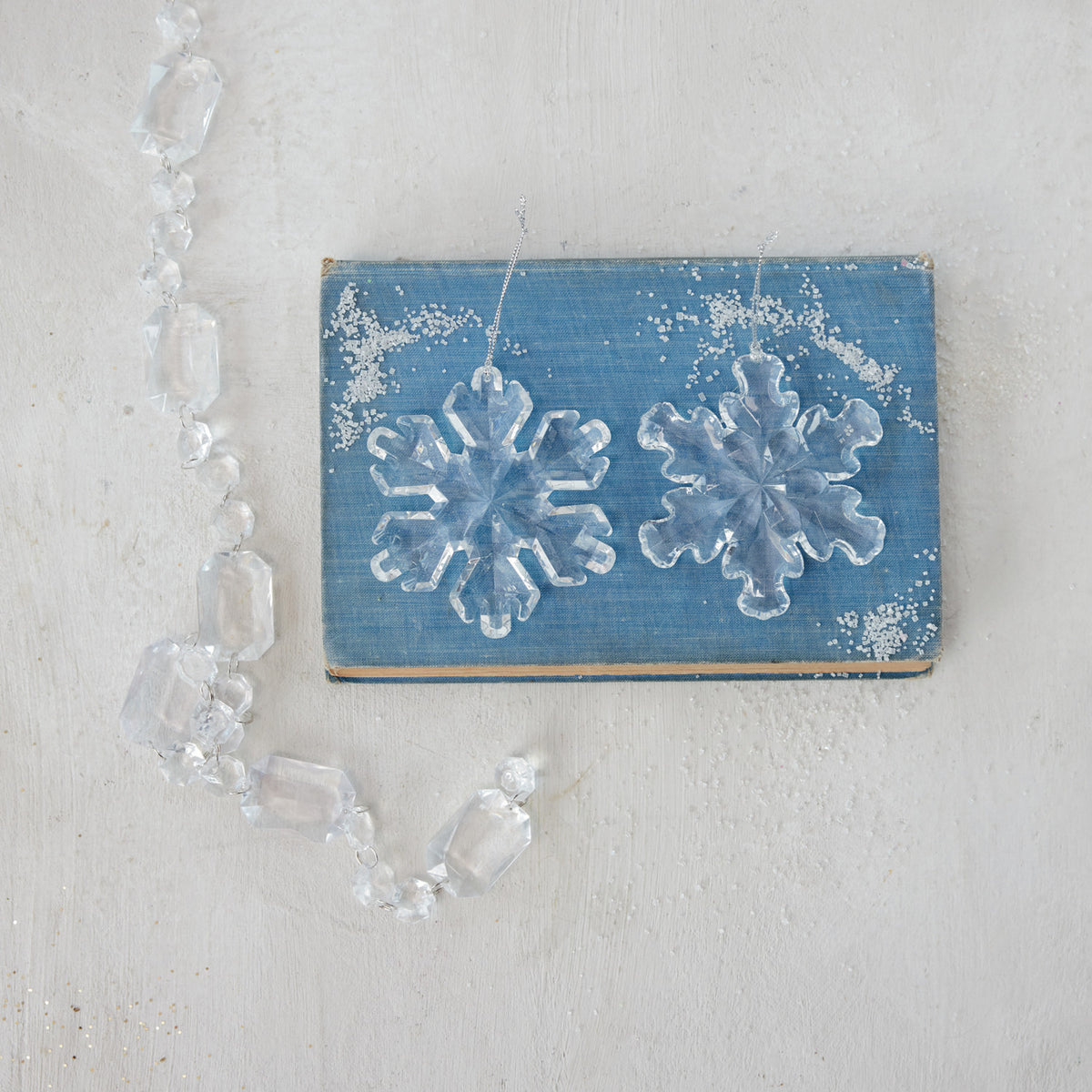 2 assorted Acrylic Snowflake Ornaments arranged on a blue book with crystal garland draped nearby.