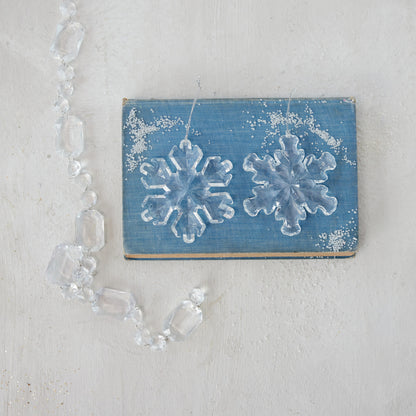 2 assorted Acrylic Snowflake Ornaments arranged on a blue book with crystal garland draped nearby.