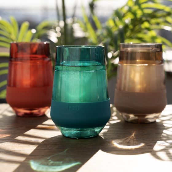 3 assorted colors of wine freeze glasses set on a porch with greenery in the background.