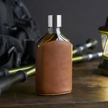 A brown leather-wrapped glass flask with a stainless steel screw-on top, displayed on a table with a flashlight and a backpack in the background.