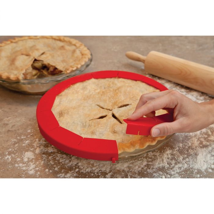 the adjustable pie shield displayed on a baked pie sitting on a flour dusted surface next to a rolling pin and pie