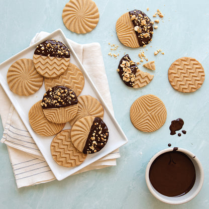 top view of coutertop with a tray of cookies half dipped in chocolate and nuts, additional cookies arranged of the tray, and a bowl of chocolate sauce.