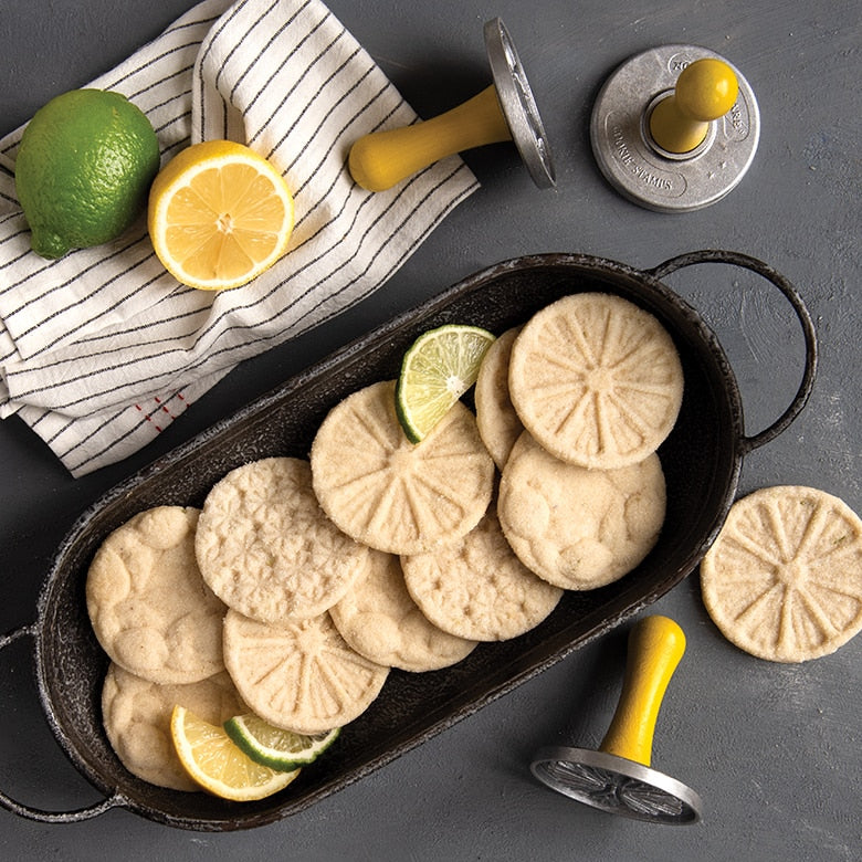 dish filled with citrus stamped cookies on a table with stamps, dishtowel, and fruits.