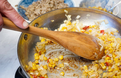 a person cooking corn and onions in a stainless steel skillet with the olive wood spoon