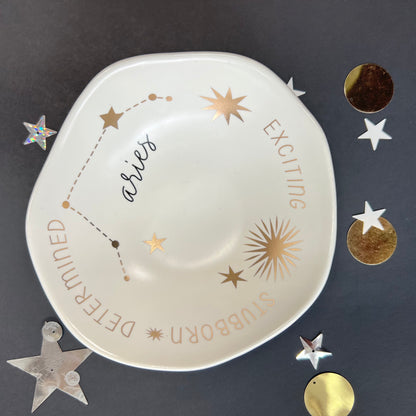cream dish with gold stars and "Aries Exciting Stubborn Determined" around the inner rim on a black background with scattered stars and orbs.