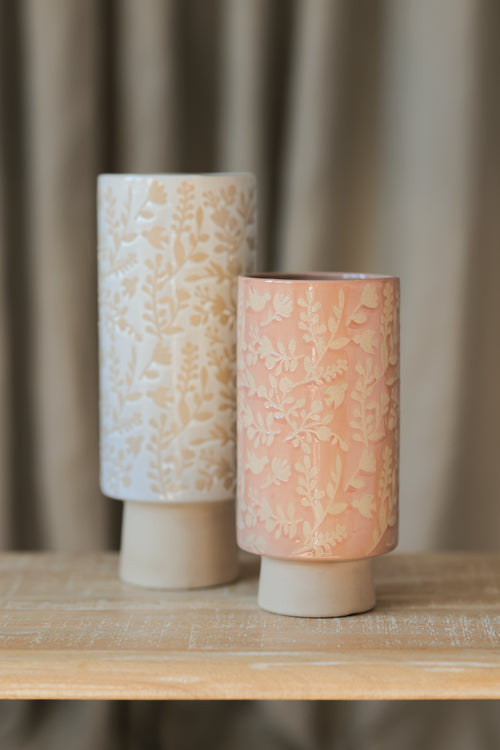 two cylindrical vases with pedestal bases on wooden table. taller vase has creamy white glaze with a floral design, shorter vase has blush pink glaze with a floral design.