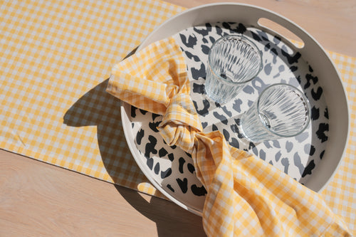 round bamboo melamine tray with handles on yellow check table runner. tray is off-white with a raised rim and cut out  handles in rim, the bottom of the tray has a black cheetah print design. The tray has the glasses and a yellow check napkin on it.