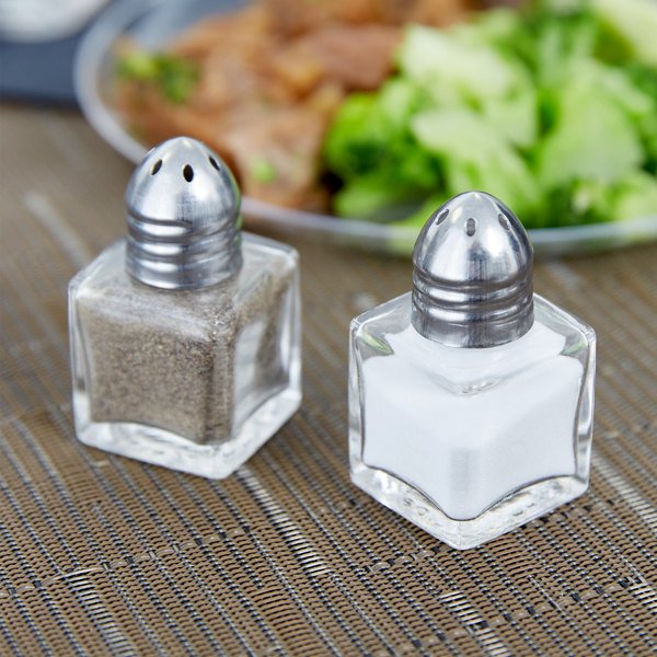 2 square shakers filled with salt or pepper on table with plate of food in background.